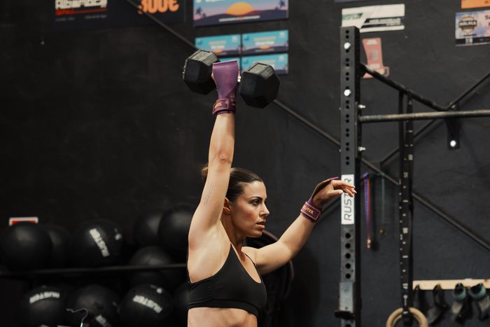 A woman pressing a dumbbell overhead in a gym, kettlebells and rigs behind her