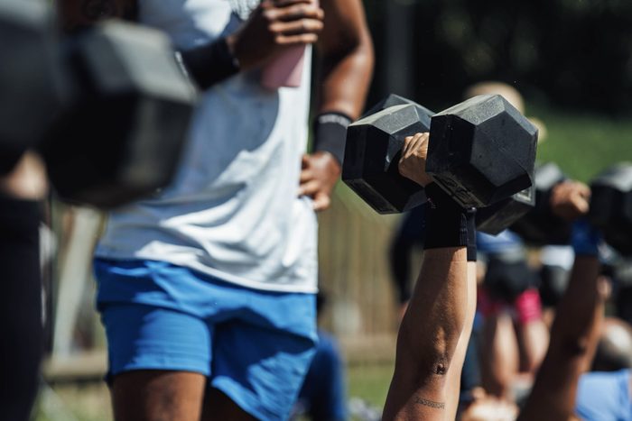 Partner dumbbell work outdoors, one athlete lying on the grass as the other stands over her