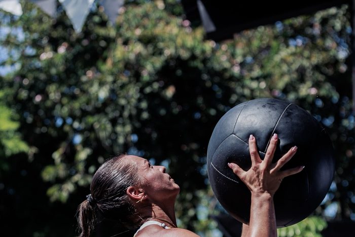 A woman reaching up to catch a heavy wall ball at an outdoor gym, trees behind