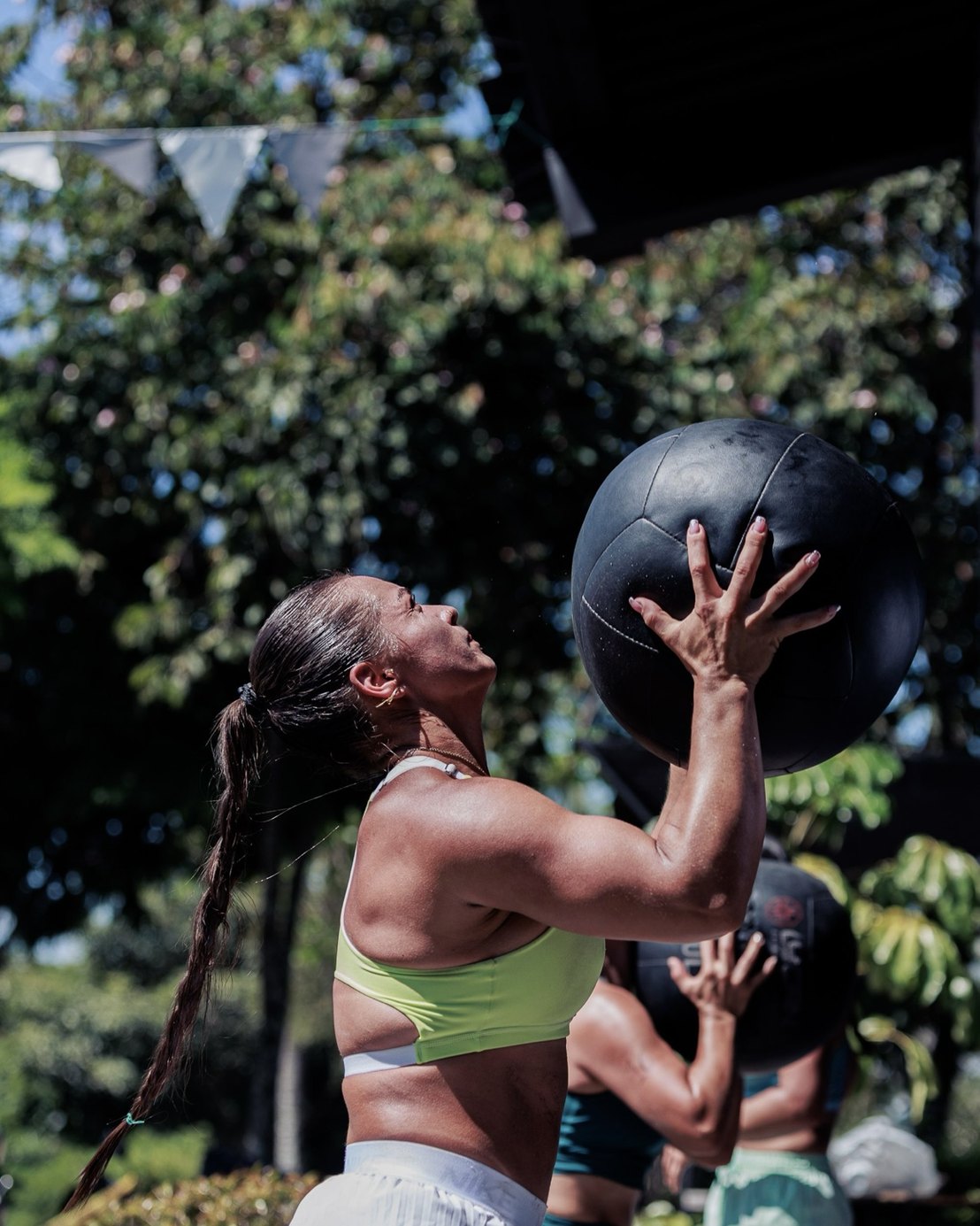 Athlete reaching up to catch a heavy wall ball at an outdoor gym, trees behind